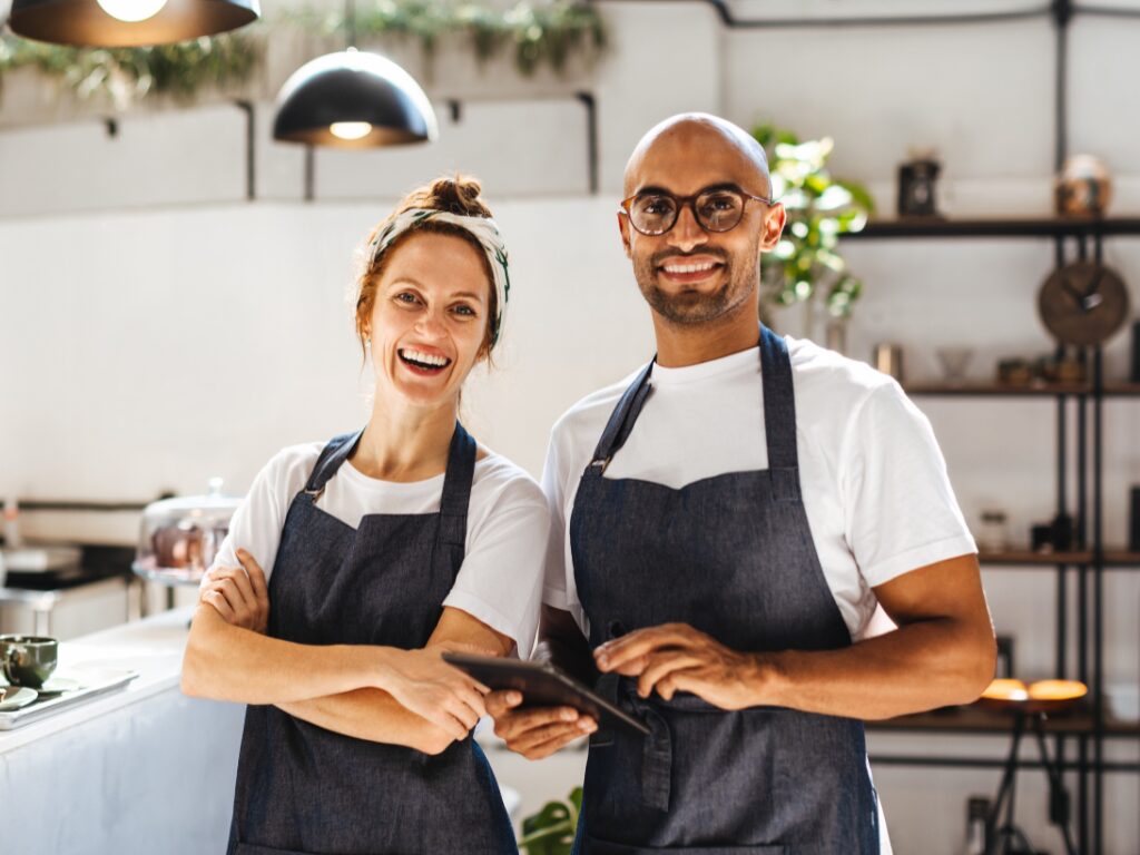 A man holding a tablet and a woman with her arms crossed smile directly at the camera, business owners wearing dark denim aprons happy because of positive working capital due to financing solutions