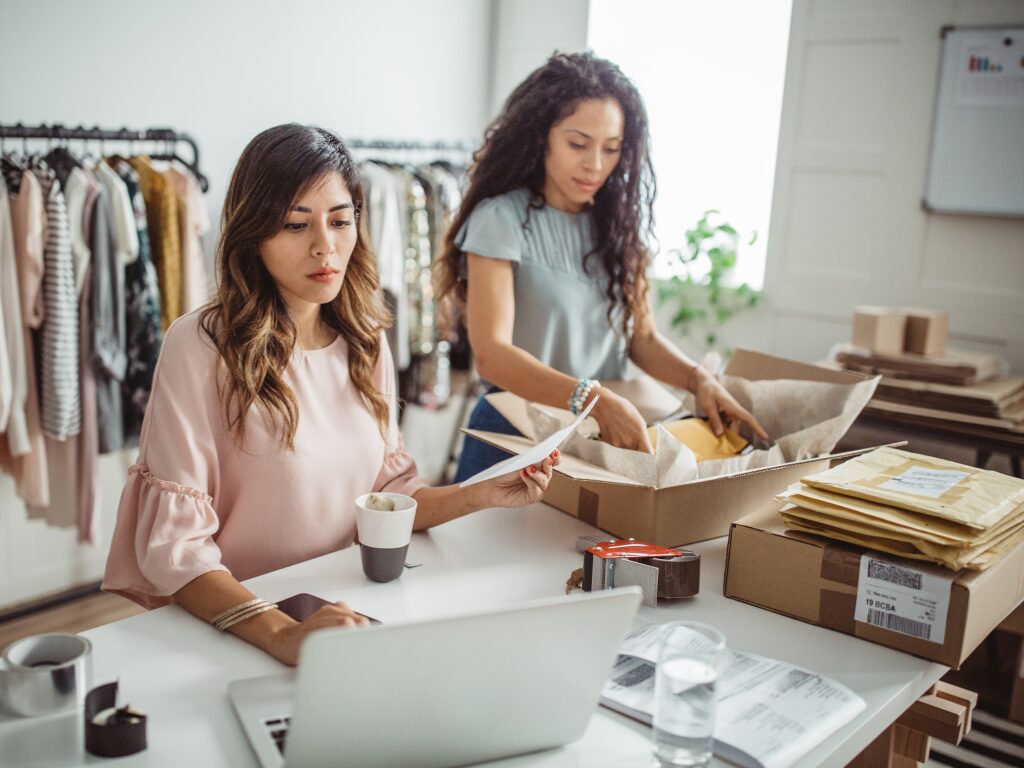 Two women working on their small business, one woman doing budgeting to ensure they have enough working capital, the other woman packing an order into a box