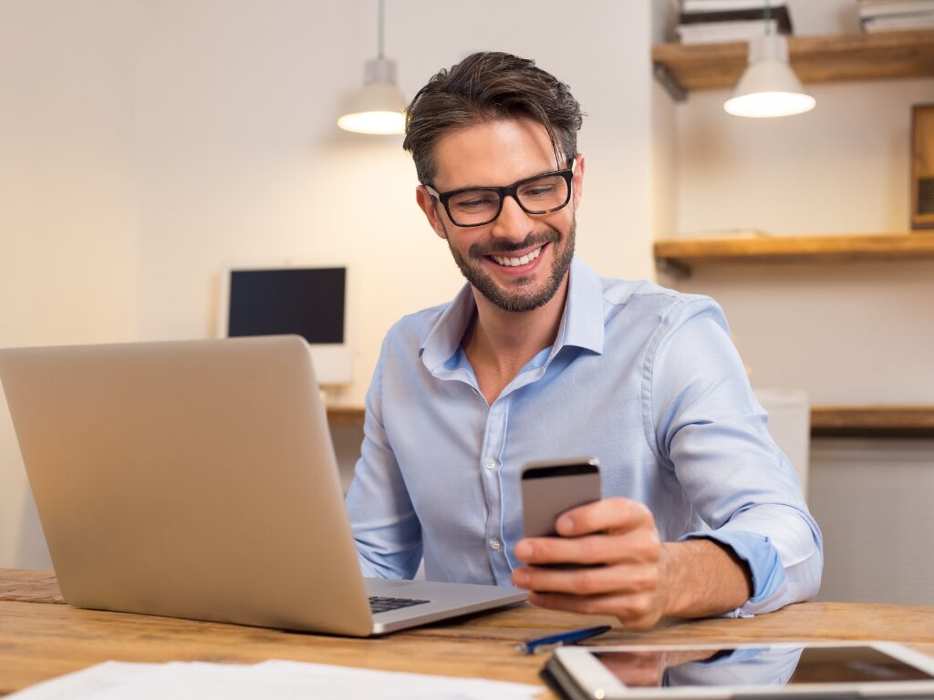 A man in front of his laptop smiles as he checks his phone, business owner learning he is qualified for a working capital loan