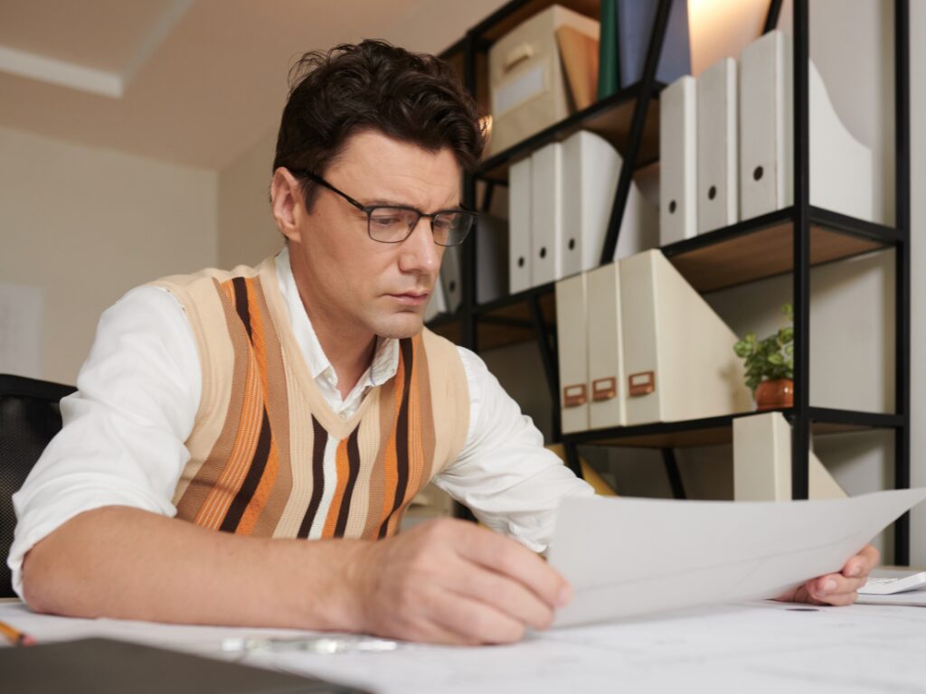 A man in glasses is seated at a desk reading a document with a slightly worried expression, business owner facing working capital funding problems