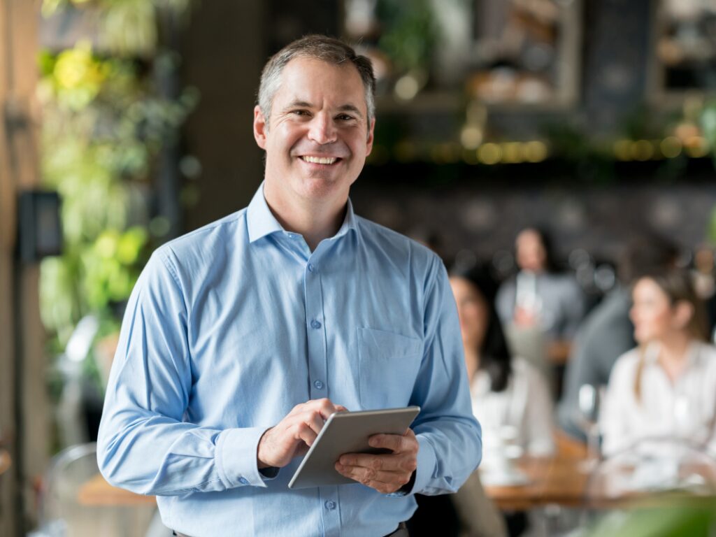 A man holding an electronic tablet smiles directly at the camera, a business owner inside his restaurant smiling, stable business because of tax debt loans