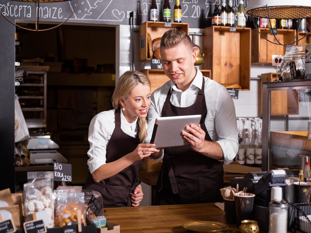 A man and a woman in aprons working behind the counter at a cafe or bakery, reading the screen of an electronic tablet together, business owners exploring working capital loan alternatives