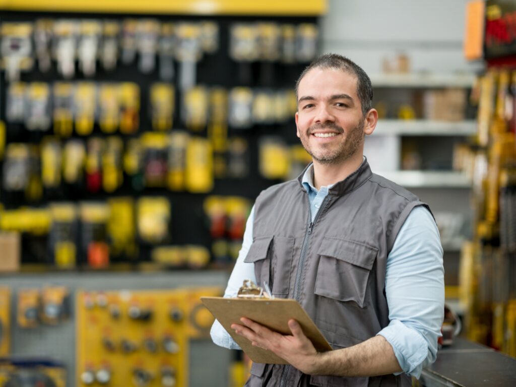 A man holding a clipboard working inside a hardware store looking directly at the camera while smiling, a business owner choosing working capital loans