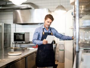 A man in an apron reads the screen of an electronic tablet while working inside an industrial kitchen, business owner looking for working capital loans alternatives