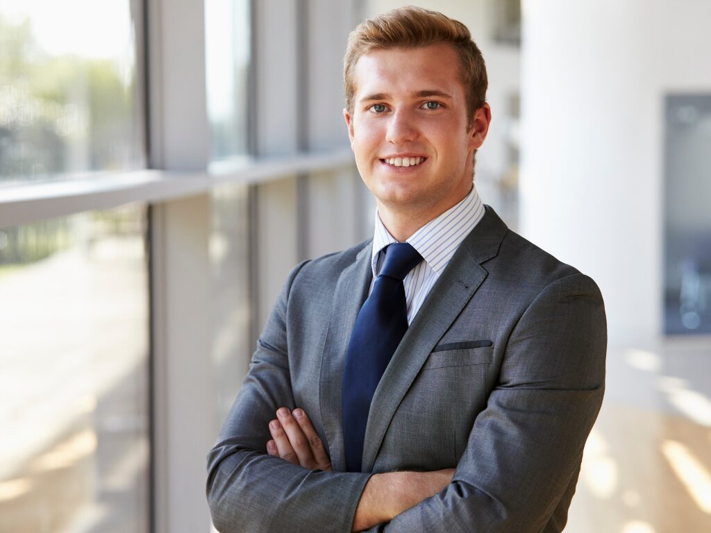 A man standing at a brightly lit professional setting smiles directly at the camera for a portrait with his arms crossed in a confident pose, a concept photo of an Australian expat seeking tax debt loan solutions