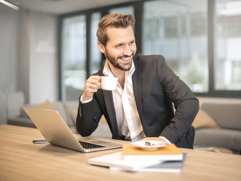 A man in a suit in a professional setting sits in front of his laptop while holding a cup of coffee and smiling off camera, an expat discussing tax debt relief options