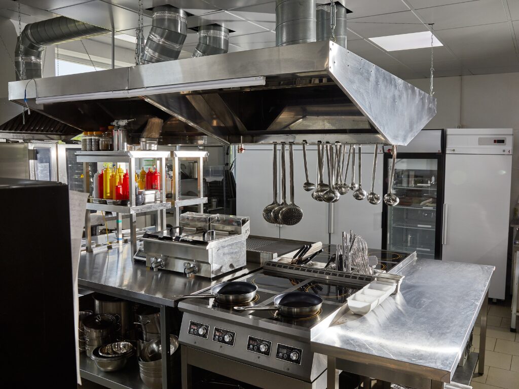 An empty fully-equipped industrial kitchen with stainless steel surfaces, fryers, range hoods, shelves, utensils, condiments, and several refrigerating units