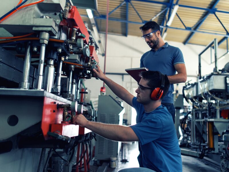 Two factory workers wearing protective gear for the eyes and ears tend to the parts of a large manufacturing machine, checking manufacturing equipment