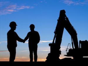 A silhouette photo showing two men in hard hats shaking hands next to an industrial excavator, construction workers or business owners shaking hands next to heavy equipment