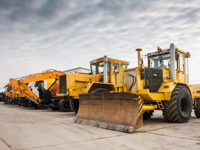 A row of different heavy equipment for construction and industrial works, yellow wheeled equipment like cranes and tractors parked next to each other with a background of grey clouds