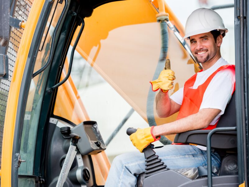 A crane operator inside the control car of his crane poses with smile and a thumbs up towards the camera