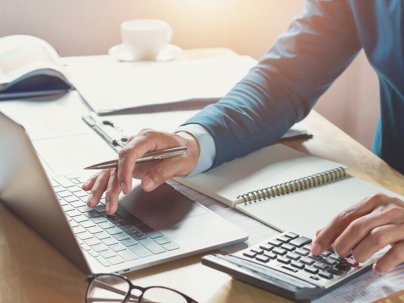 Cropped photo of a man using his laptop and calculator while holding a pen, calculating taxes and tax to avoid going into tax debt