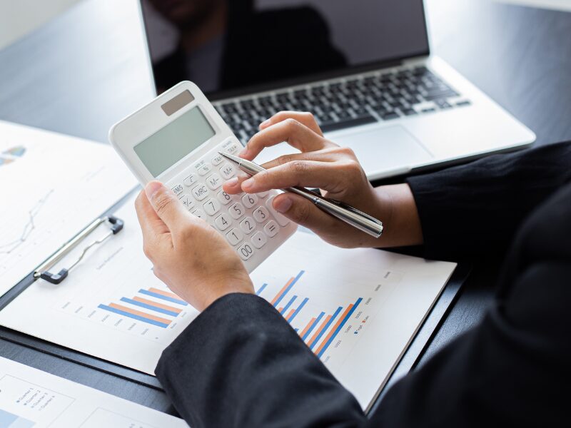 Cropped photo of a person holding a pen while using a white calculator, documents and a laptop visible in the background, an investor calculating the cost of a residential loan