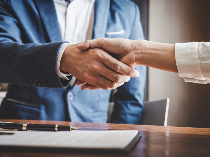 Cropped photo of a man in a blue suit shaking hands with a person, a pen and a contract visible on the table, an investor and lender agreeing on terms for an investment loan