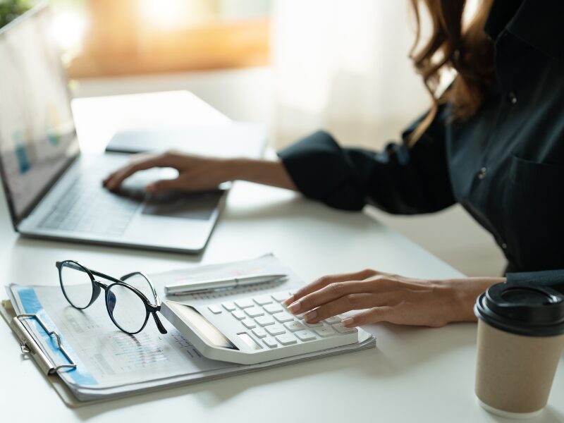 A woman uses a calculator and her laptop at the same time, a paper cup of coffee, glasses, documents, and a pen on top of the table, investor calculating the cost of a loan and the potential gains they can get