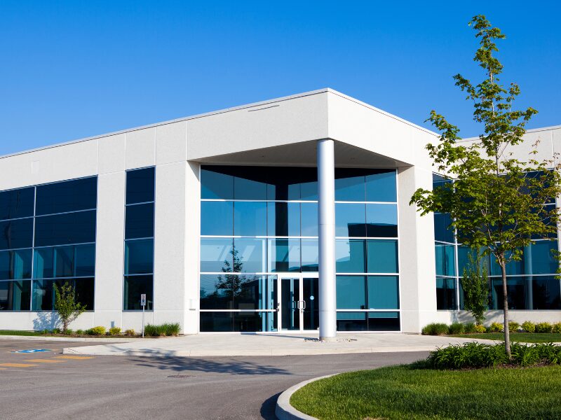 Exterior of a low-rise white commercial building with large bluish tinted windows, with a raised area in front with a small tree, shrubs, and grass