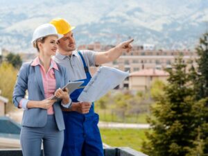 A builder holds up plans and points forward to something off camera while a developer looks on and smiles, a man and a woman in hard hats overlooking a construction site