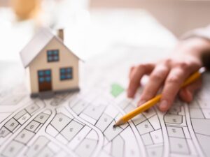 A hand holding a pencil points to a specific lot in a property development plan for a suburb, a miniature model house sits on top of the plan