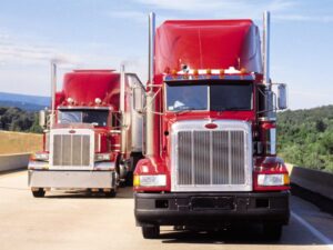 Two bright red freight trucks driving side by side with the one on the left slightly behind, two large transport trucks driving down the road