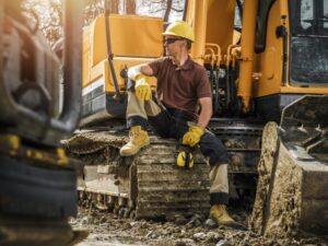 A heavy equipment operator sits atop the wheels of his equipment with one leg raised up and looking to the left, a business owner choosing between equipment leasing vs equipment loans