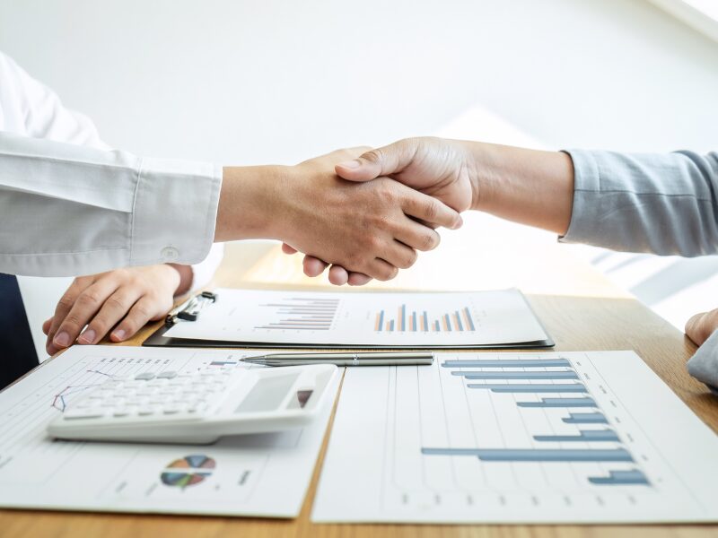 Cropped photo of two people shaking hands, with documents, a pen, and a calculator visible on the table below, borrower and lender agreeing on terms for a debt consolidation loan