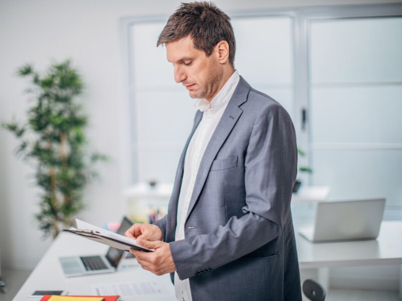 A man in a professional setting reads documents on a clipboard, business owner reading through his current debts for consolidation