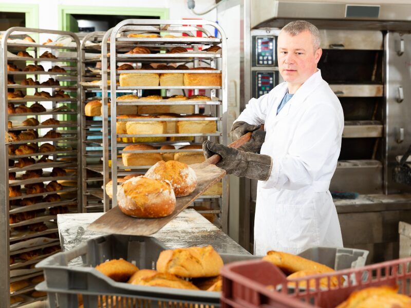 A baker in a commercial bakery handles freshly baked bread using a wooden peel, baker surrounded by racks of bread, with a large oven behind him