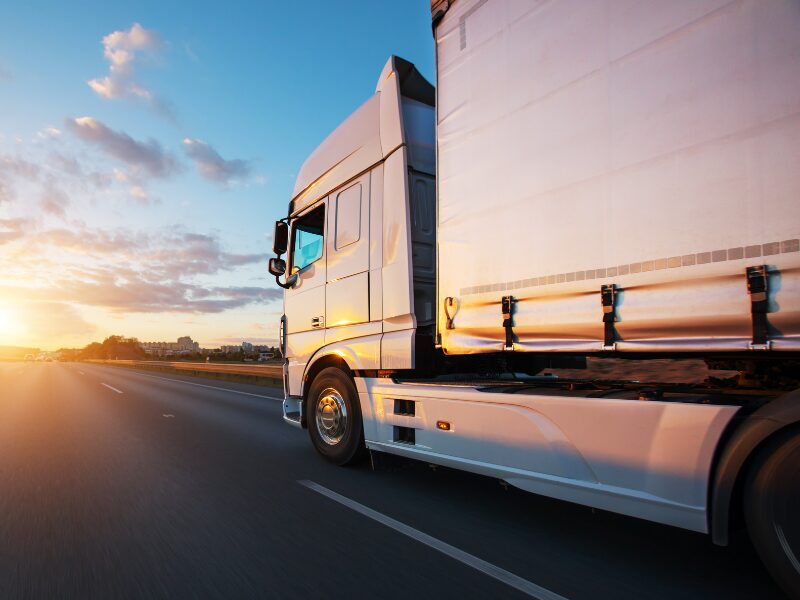 Side view of a white truck driving down a major road with the sunrise ahead in the horizon