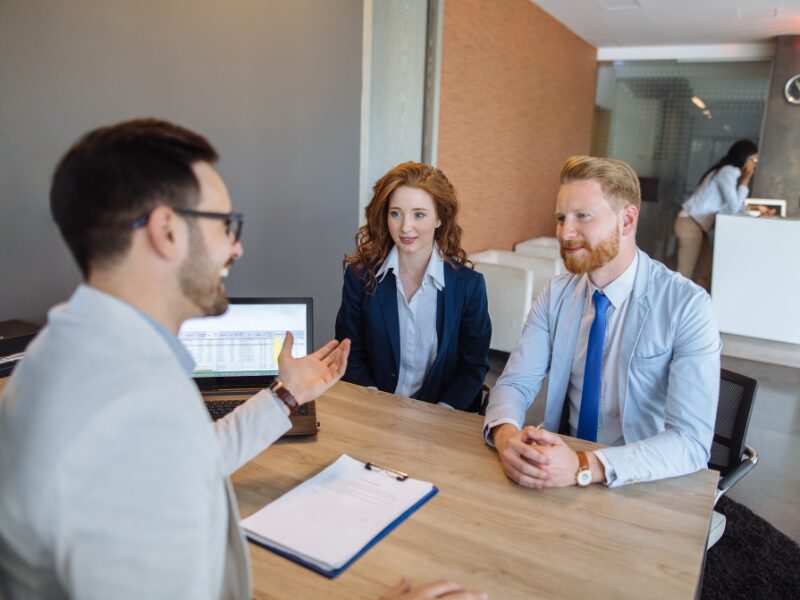 A man and a woman in a professional setting sit across a lender or a lending expert, seeking advice for refinancing a caveat loan
