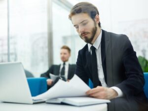 A man in a suit reads documents with a serious expression, another man in a suit sitting far behind him, men in a professional setting
