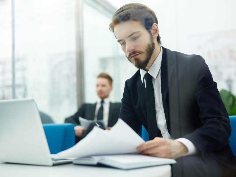 A man in a suit reads documents with a serious expression, another man in a suit sitting far behind him, men in a professional setting