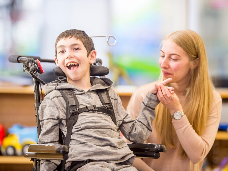 A happy child with a disability sits on a specialised wheelchair as a worker or NDIS provider holds his hand happily