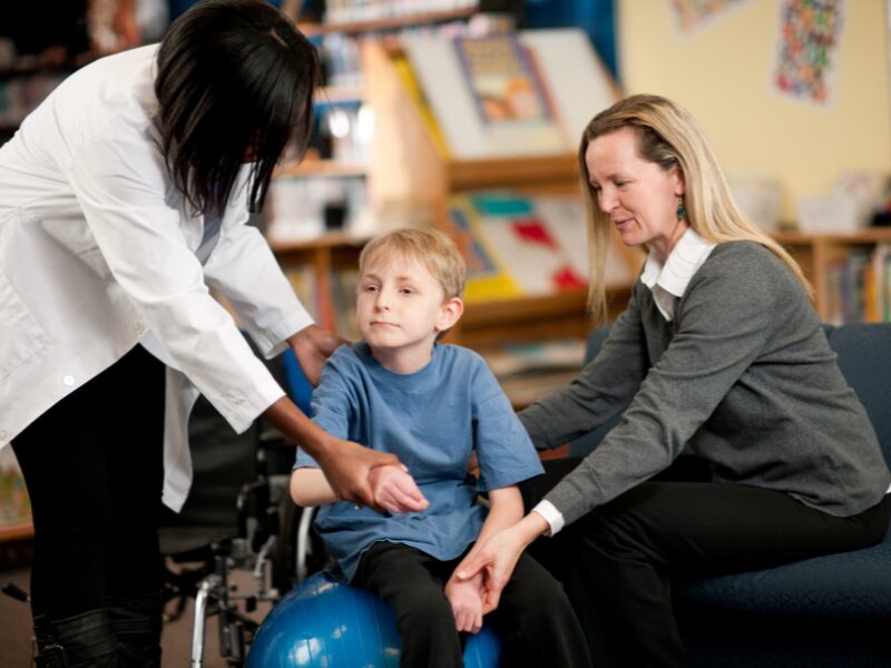 Two women assist a child with disability during physical therapy, NDIS providers assisting a participant