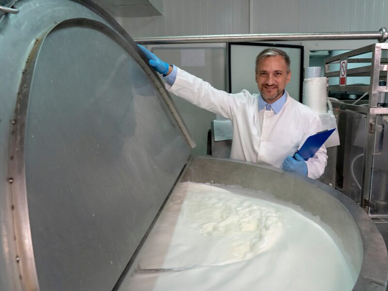 A food manufacturing business owner or worker opens a vat of boiling dairy, worker in sanitary clothing carrying a clipboard and smiling
