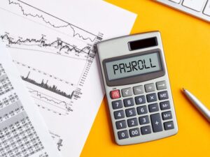 A calculator displaying the word “payroll”, documents, and a silver pen arranged on top of a bright mustard yellow background surface