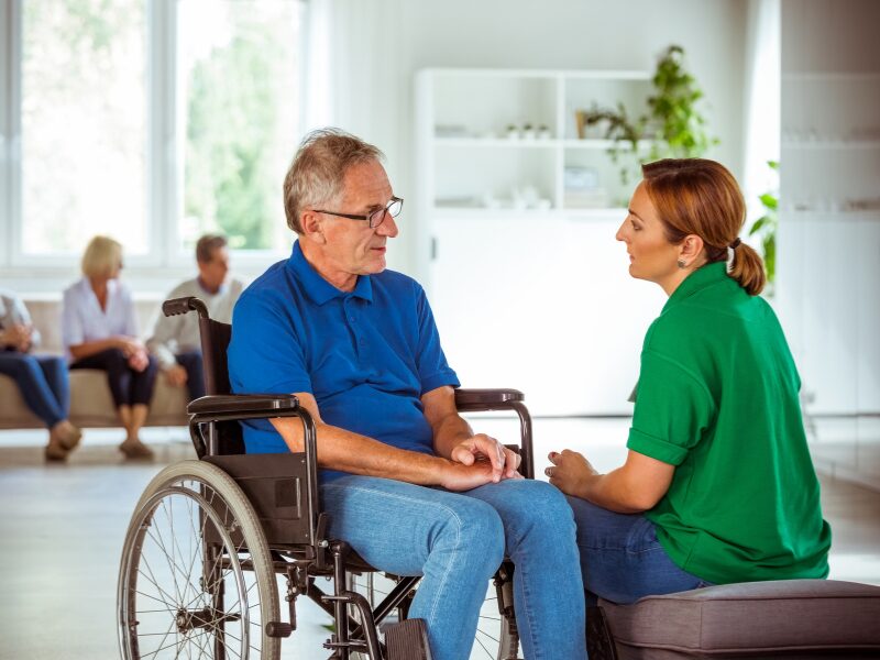 An NDIS worker sitting and engaged in conversation with an elderly man in a wheelchair, a Support worker talking to a patient or client