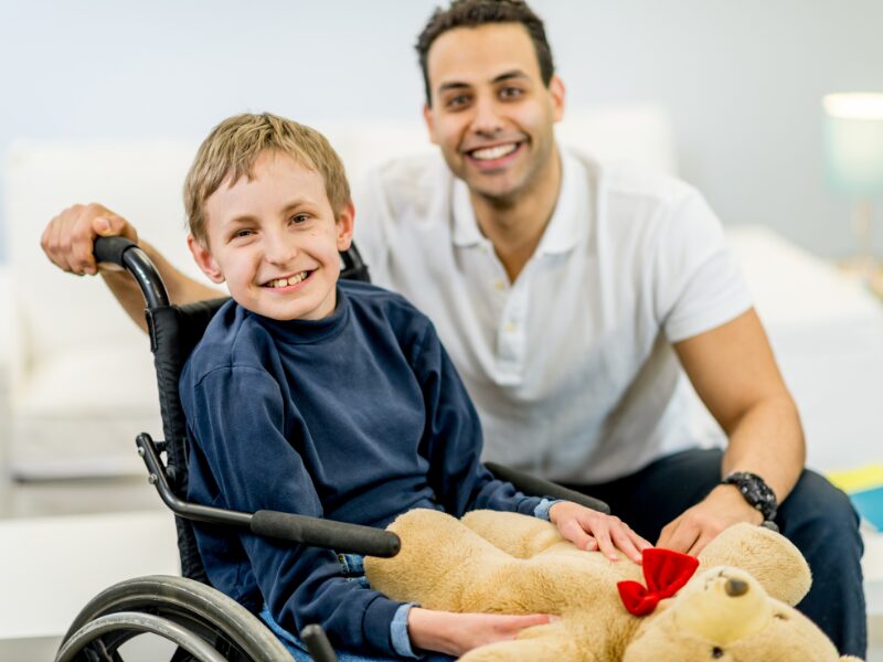 An NDIS provider smiles and poses with a child with disability sitting in a wheelchair holding a brown teddy bear with a red bow, a happy NDIS worker with a client