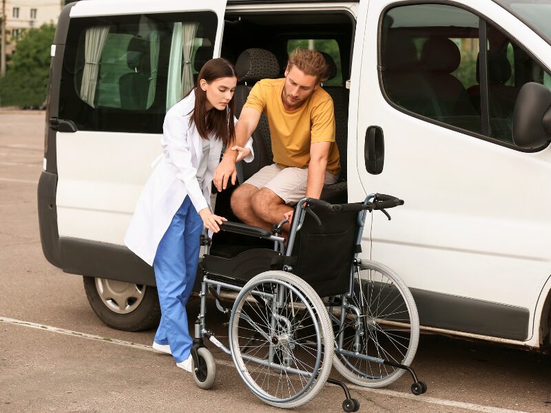 A healthcare or NDIS worker in a white coat and scrubs helps a man with disability get down from a white van and onto a wheelchair