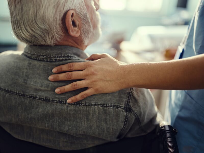 An NDIS provider provides comfort to an elderly participant on a wheelchair by gently placing her hand on his shoulder