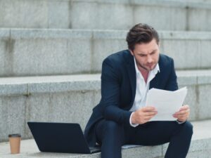 A man in a suit sits on the steps of an outdoor staircase reviewing documents with a stressed expression, a business owner reviewing their general interest charges from the ATO