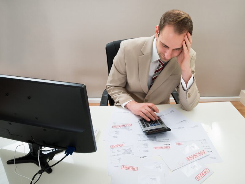 A stressed man uses his calculator while his table is littered with overdue notices from various debts, including tax debt