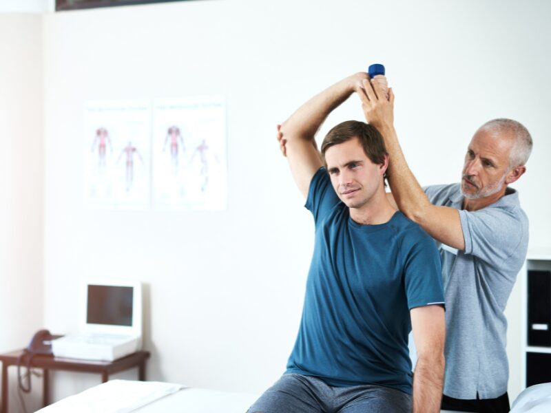 A therapist works with a client as he does arm stretches while holding a small dumbbell