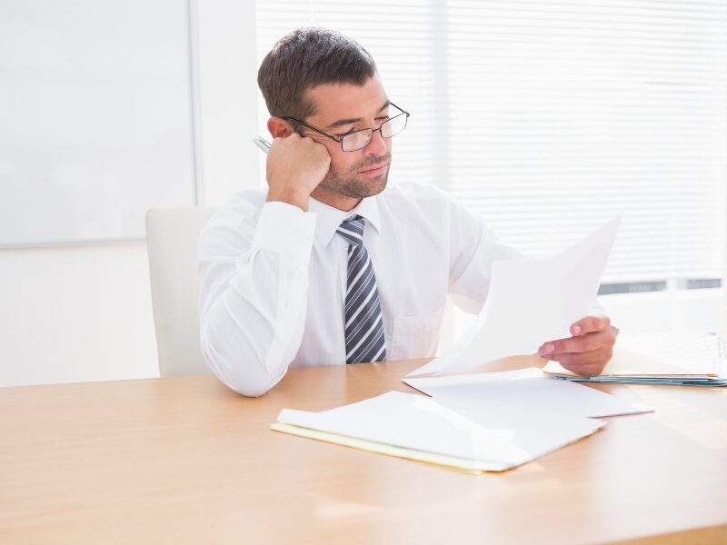 A man in a professional setting reads a document while resting his head on his hand, reading about SIC on unpaid business taxes