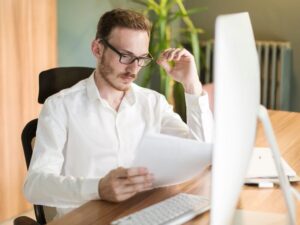 A man seated in front of his computer reads documents with a serious expression, a business owner reading an amended assessment notice from the ATO