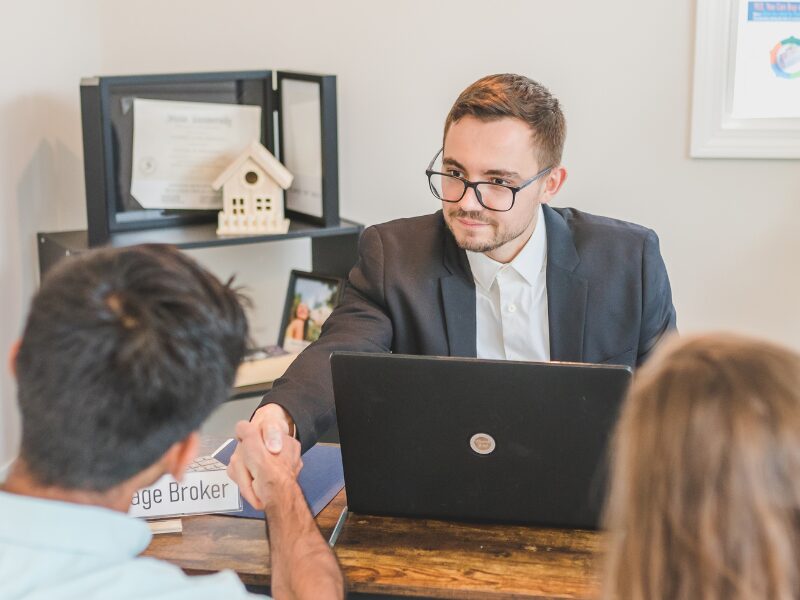 A man seated next to a woman shakes the hand of a mortgage broker seated across them, borrowers agreeing to a home loan with a mortgage offset account feature