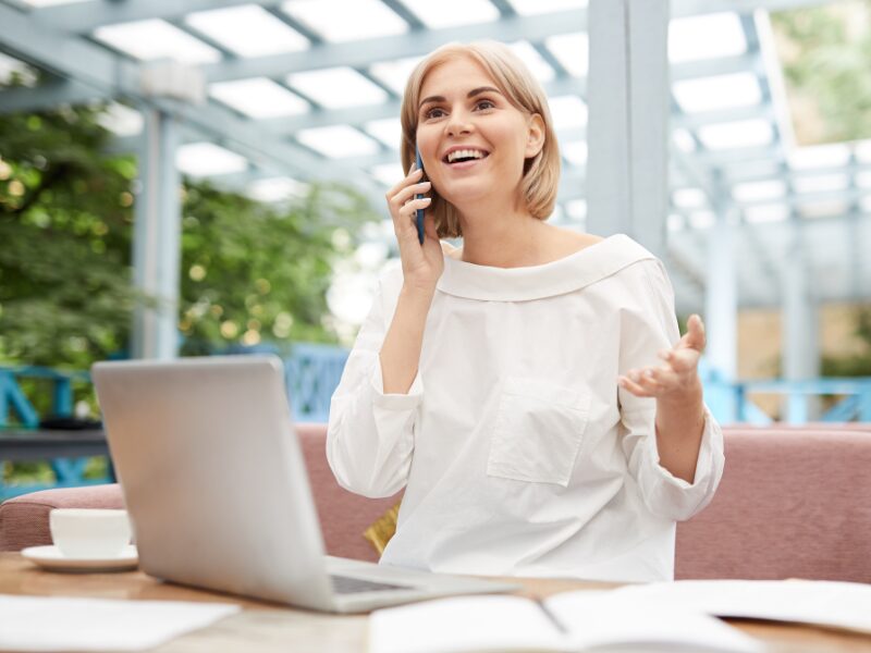 Cheerful woman on the phone sitting at a restaurant or cafe outdoors, laptop, notebook, and coffee cup on the table, home owner discussing offset accounts with their broker or lender