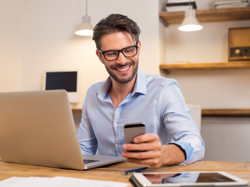 A man in glasses smiles as he looks at his phone, an electronic tablet and a laptop on the table, man happy about interest savings on his loan