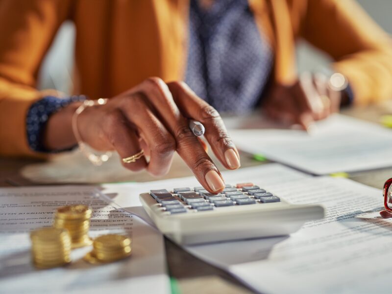 Cropped photo of a woman using a calculator, documents and piles of coins visible on the table, borrower calculating the interest savings brought by extra repayments