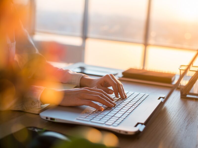 A person’s hands typing on a laptop computer, sunlight entering the window and illuminating the room, a person making online extra payments on their loan