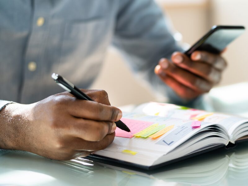 A man holds his phone while he writes on a planner, business owner marking down the date of the policy change to ensure he pays tax debts on time